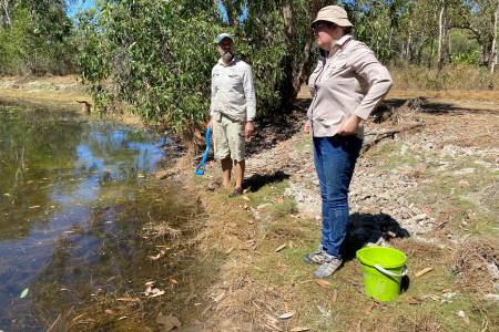Two people standing on the bank of a water body with a green bucket in the foreground and trees in the background