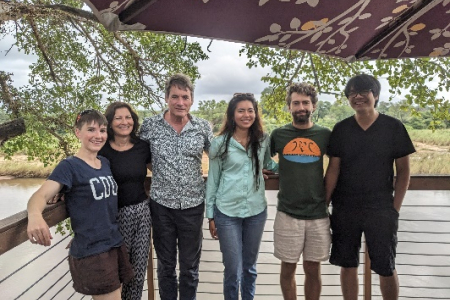 six people standing in a row, posing for photo, with vegetation in background