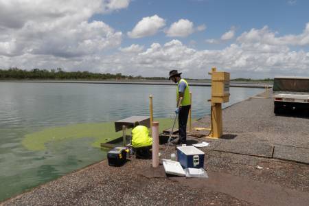 two people, one standing, one squatting, on concrete edge of large pond, collecting samples of water near a patch of greenish algae