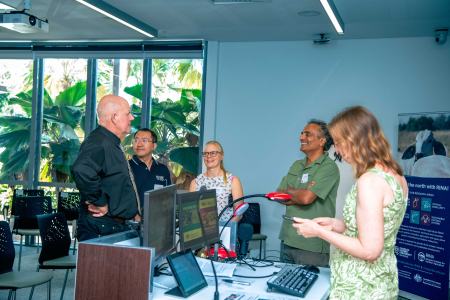 Five people stand chatting near lecture room lectern, with large palm fronds visible through windows in the background