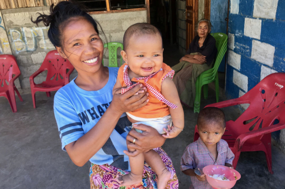 A woman holding a small child with another child standing beside her and an old woman sitting in the background
