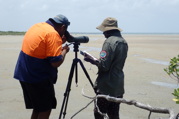 two men on a beach, one looking through a telescope, one writing in a notebook
