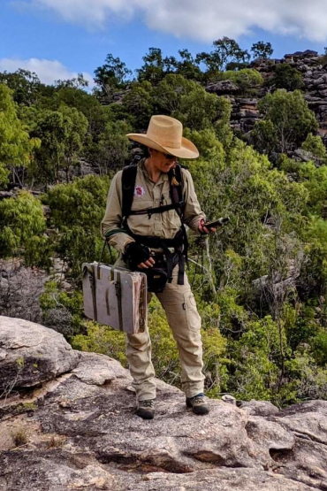Person wearing hat standing on rocks looking at GPS, with trees in background