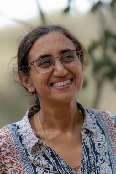 Kamaljit Sangha, head and shoulders, wearing a patterned top and glasses, with a blurry green and brown background